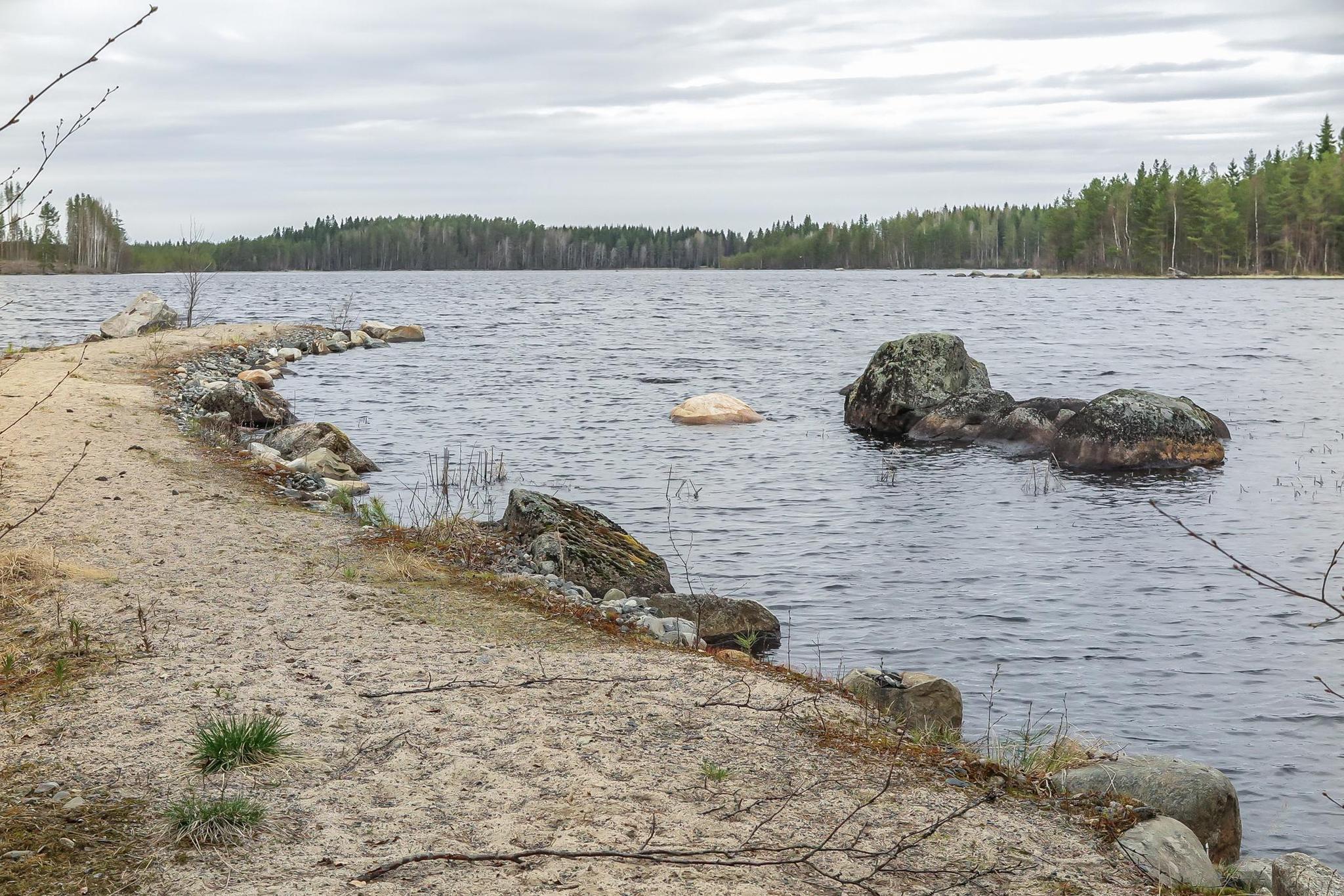 Scenic view of a property featuring two cabins on a narrow peninsula by Lake Saarivesi, with a shallow, sandy beach and private dock. The main cabin, built in 1995, includes a covered porch and separate sleeping quarters. Adjacent is a sauna cabin built in 2012, with an upper sleeping loft and a sauna accessible by steep stairs. Amenities include a grill shelter, cold cellar, and a rowboat. The lush, tree-lined surroundings emphasize a serene, secluded retreat ideal for relaxation and enjoying nature